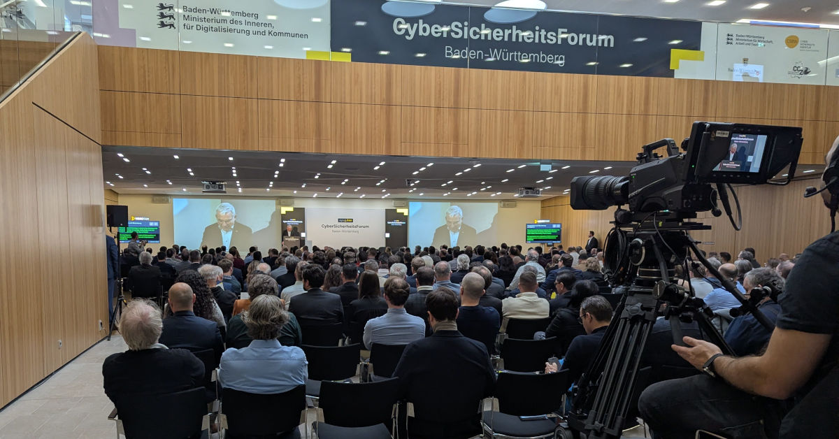 Conference hall with many attendees seated, large screens on stage showing the opening session of the CyberSicherheitsForum Baden-Württemberg.