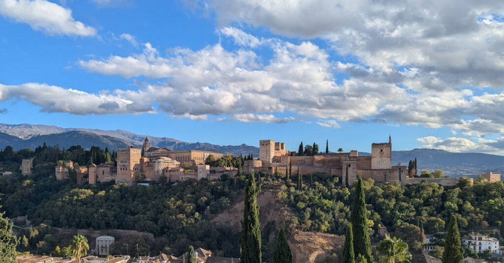 Panoramic view of the Alhambra on a wooded hill above Granada, with cypress trees in the foreground and mountains under a sky of scattered clouds.